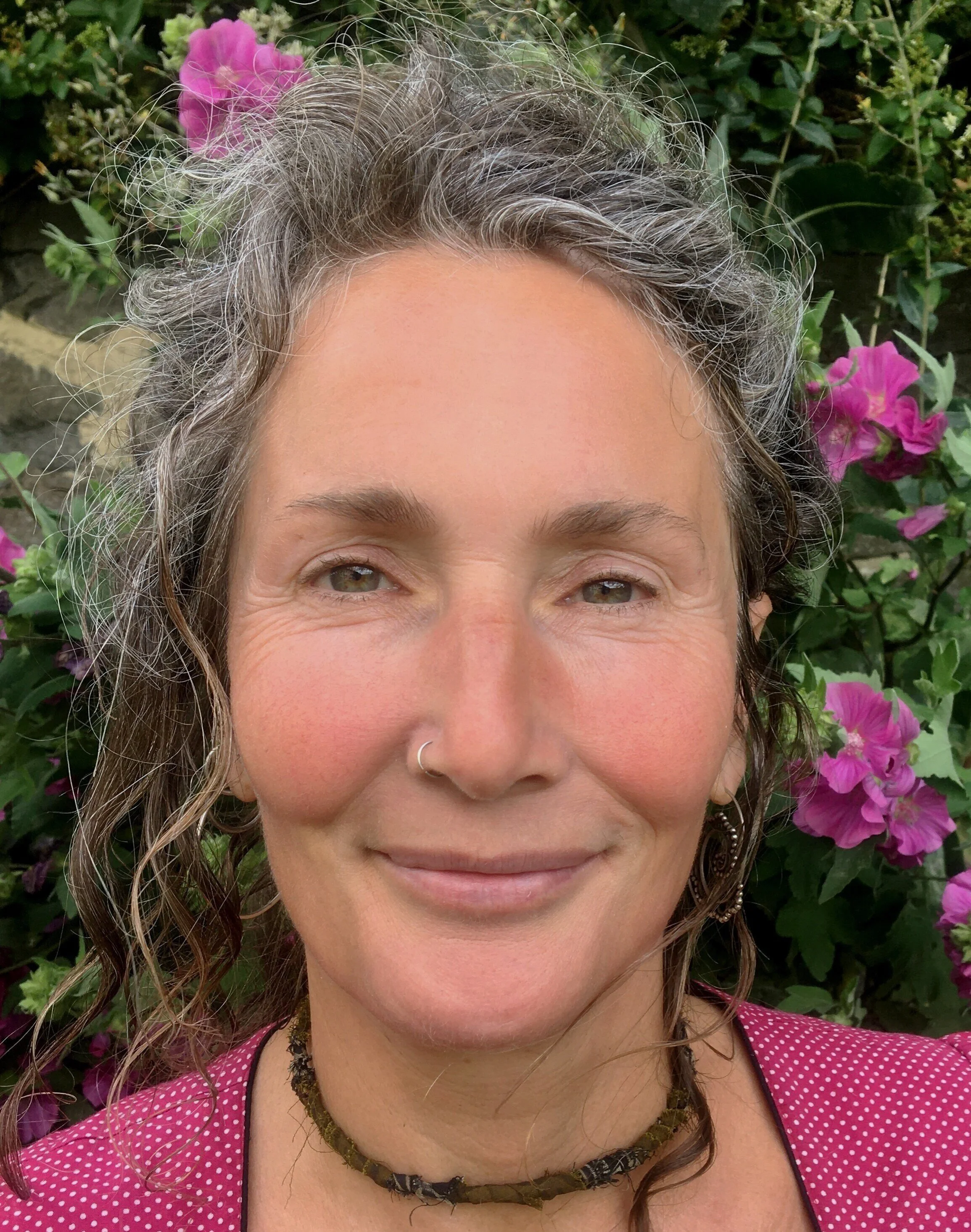 A smiling doula stands in a garden with pink flowers in the background.